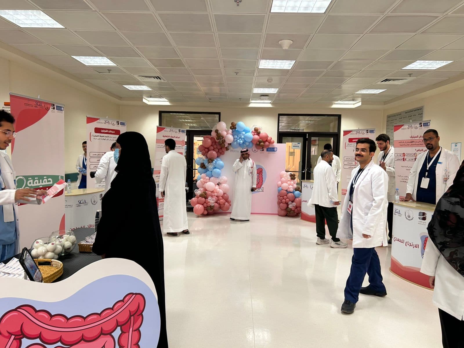 Wide view of the KKUH outpatient hall during the campaign — themed booths for IBD, colorectal cancer, GERD, and IBS arranged beneath a pink-and-blue balloon arch.